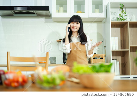 Asian woman chats on the phone and drinks coffee in a cozy kitchen with fresh vegetables. A vibrant lifestyle scene perfect for food, wellness, and communication concepts. Asian woman chats on the phone and drinks coffee in a cozy kitchen with fresh vegetables. A vibrant lifestyle scene perfect for food, wellness, and communication concepts. 134865690