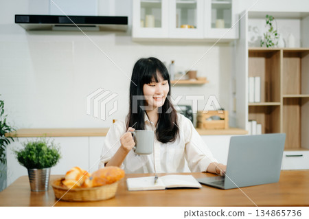 Asian woman working remotely on a laptop and tablet in a cozy kitchen workspace. Perfect for education, freelance Asian woman working remotely on a laptop and tablet in a cozy kitchen workspace. Perfect for education, freelance 134865736