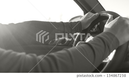 Hands gripping steering wheel of a car while driving under bright sunlight, showcasing focus and determination on the road ahead, toned with Colour of the year Cloud Dancer. Hands gripping steering wheel of a car while driving under bright sunlight, showcasing focus and determination on the road ahead, toned with Colour of the year Cloud Dancer. 134867238