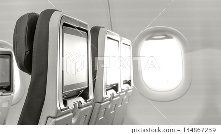 Airplane interior featuring empty passenger seats with personal screens and a bright window, creating a serene travel atmosphere with copy space, toned with Colour of the year Cloud Dancer. Airplane interior featuring empty passenger seats with personal screens and a bright window, creating a serene travel atmosphere with copy space, toned with Colour of the year Cloud Dancer. 134867239