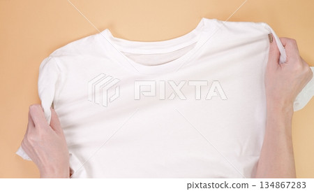 Hands Preparing a Clean White T-Shirt on Beige Background, Ready for Custom Design or Text Hands Preparing a Clean White T-Shirt on Beige Background, Ready for Custom Design or Text 134867283