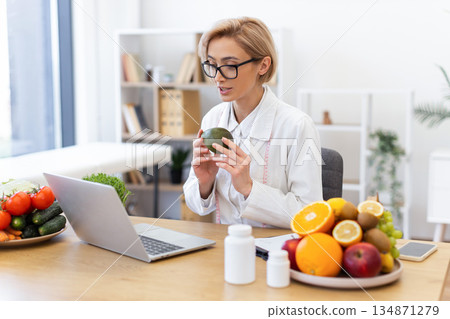 A nutritionist consults online, holding an avocado and surrounded by fresh fruits and vegetables, promoting healthy eating habits. A nutritionist consults online, holding an avocado and surrounded by fresh fruits and vegetables, promoting healthy eating habits. 134871279