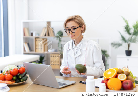 A dietitian consults with a client online, holding an avocado and surrounded by fresh fruits and vegetables. 134871280