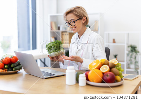 A smiling nutritionist in a white coat holds sprouts, discussing nutrition with a laptop and a table full of fruits and vegetables. A smiling nutritionist in a white coat holds sprouts, discussing nutrition with a laptop and a table full of fruits and vegetables. 134871281