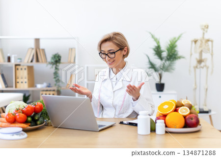 A smiling dietitian consults with a patient online, surrounded by healthy food and supplements. A smiling dietitian consults with a patient online, surrounded by healthy food and supplements. 134871288