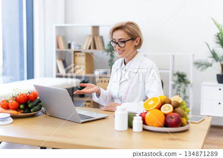 A dietitian in a white coat consults with a patient online, surrounded by fresh fruits and vegetables. 134871289