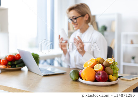 A nutritionist consults with a client online, surrounded by fresh fruits and vegetables, promoting healthy eating habits. 134871290