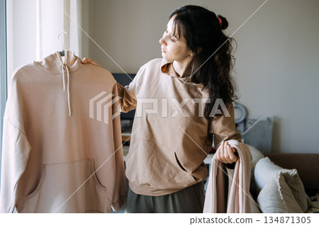 Woman holds hoodie and trousers while standing in living room. Hybrid work style, domestic comfort, functional wardrobe. 134871305