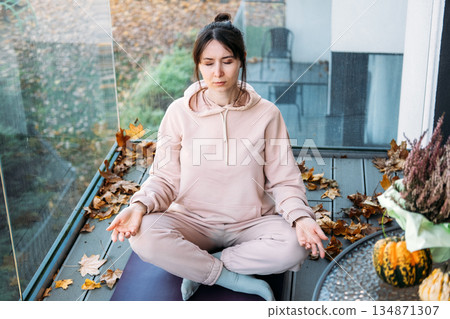Woman sits in meditative pose on balcony with autumn leaves and seasonal decor. Traditional yoga, eastern philosophy, meditation practice, authentic wellness. Woman sits in meditative pose on balcony with autumn leaves and seasonal decor. Traditional yoga, eastern philosophy, meditation practice, authentic wellness. 134871307
