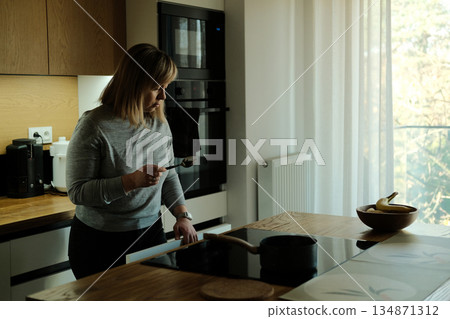 Woman stands in modern kitchen holding spoon while looking down at induction stove with pot. Induction cooktop, energy-saving appliances, sustainable kitchen, eco-friendly home. 134871312