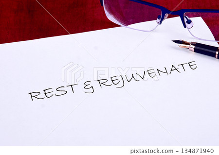 A crisp white paper displays the phrase Rest and Rejuvenate, accompanied by sophisticated spectacles and a fountain pen on a rich, crimson backdrop in the afternoon 134871940