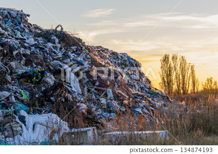 Piles of garbage are seen at a landfill site with a backdrop of trees and a sunset sky in the evening hours 134874193