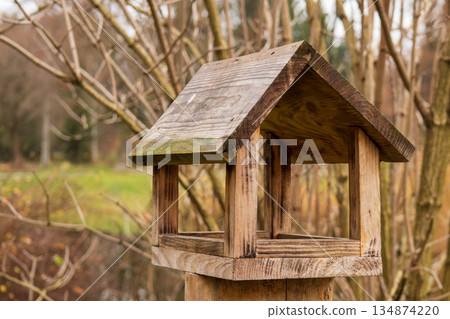A wooden birdhouse is placed on a post in a garden. Trees and grass surround it in an outdoor area during autumn 134874220