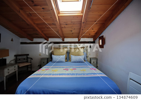 Cozy attic bedroom with blue bedding under a wooden sloped ceiling and skylight 134874609
