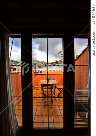Cozy rooftop terrace with orange walls, wooden table and chairs, viewed through glass doors to a cloudy sky 134874639