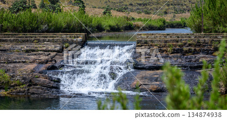 A gentle tiered waterfall cascades over dark rock into a calm pool surrounded by highland shrubs and open grassland in Horton Plains National Park. 134874938
