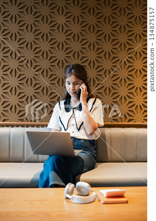 A young woman multitasks by working on a laptop and talking on the phone in a contemporary cafe setting. 134875157