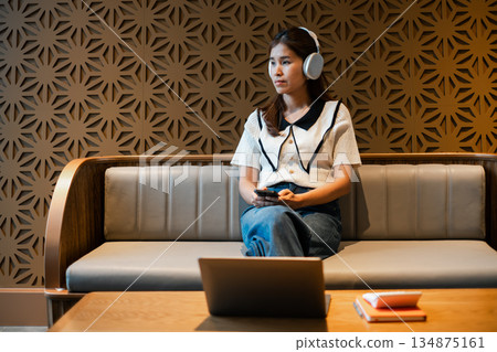 Woman sitting on a sofa with headphones, holding a smartphone, in a modern lounge with a geometric wall and laptop on the table. Woman sitting on a sofa with headphones, holding a smartphone, in a modern lounge with a geometric wall and laptop on the table. 134875161