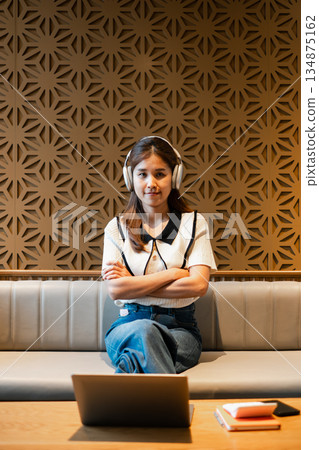 Woman wearing headphones sitting on a sofa with a laptop, set against a modern geometric wall design. Woman wearing headphones sitting on a sofa with a laptop, set against a modern geometric wall design. 134875162