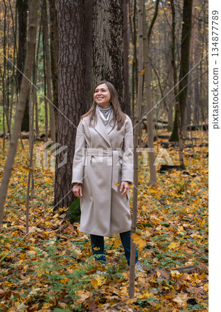 Woman in a light beige coat stands in an autumn forest, looking up among tall trees and yellow fallen leaves, calm and reflective mood Woman in a light beige coat stands in an autumn forest, looking up among tall trees and yellow fallen leaves, calm and reflective mood 134877789