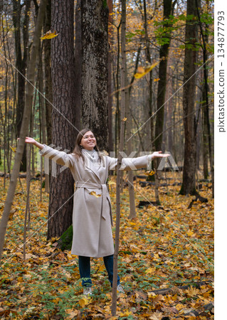 Woman in beige coat tosses autumn leaves in a forest, standing among tall trees with yellow foliage, capturing motion, freedom, seasonal outdoor mood Woman in beige coat tosses autumn leaves in a forest, standing among tall trees with yellow foliage, capturing motion, freedom, seasonal outdoor mood 134877793