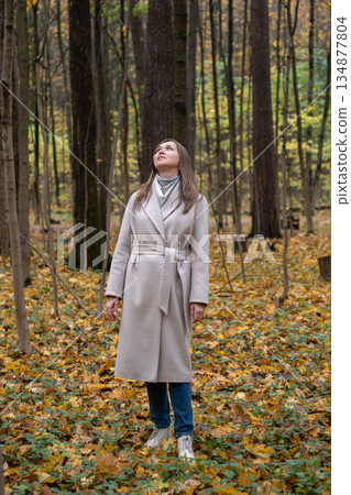 Woman in a light beige coat stands in an autumn forest, looking up among tall trees and yellow fallen leaves, calm and reflective mood 134877804