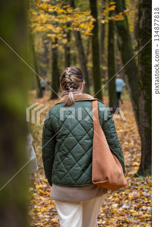 Woman with braided hair walks away along leaf-covered path in Timiryazevsky Park, Moscow, carrying brown bag, autumn forest mood Woman with braided hair walks away along leaf-covered path in Timiryazevsky Park, Moscow, carrying brown bag, autumn forest mood 134877818