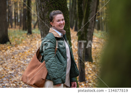 Woman in green quilted jacket leans against mossy tree in Timiryazevsky Park, Moscow, surrounded by golden autumn leaves, forest atmosphere Woman in green quilted jacket leans against mossy tree in Timiryazevsky Park, Moscow, surrounded by golden autumn leaves, forest atmosphere 134877819