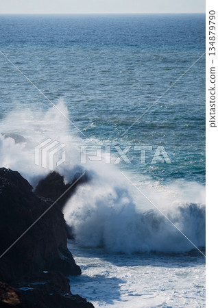 Crashing waves collide with jagged rocks along the blue ocean coastline, creating dramatic spray and foam Crashing waves collide with jagged rocks along the blue ocean coastline, creating dramatic spray and foam 134879790