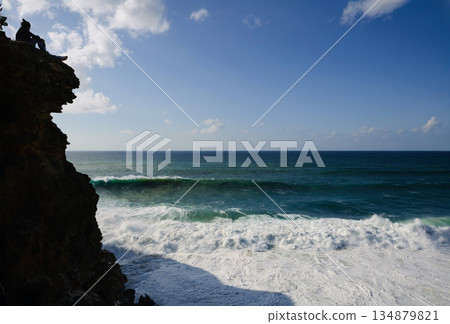 Person sits on a rocky cliff overlooking the ocean waves under a blue sky at the coast 134879821