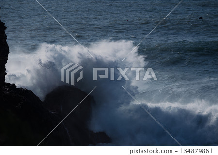 Waves crash against dark rocks along a rugged coast, creating dramatic ocean spray 134879861