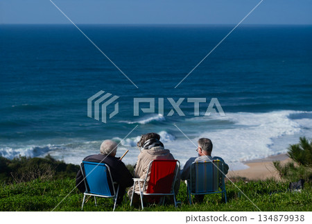 Three seniors sit on lawn chairs overlooking the sea, enjoying a peaceful coastal view 134879938