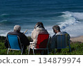 Three elderly friends sit in colorful beach chairs facing the ocean on a sunny day 134879974