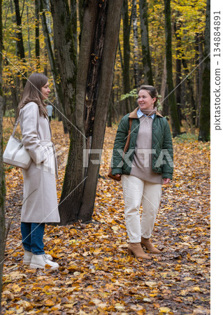 Two women walk and talk on leaf-covered path among tall trees in Timiryazevsky Park, Moscow, calm and friendly mood, natural light Two women walk and talk on leaf-covered path among tall trees in Timiryazevsky Park, Moscow, calm and friendly mood, natural light 134884891