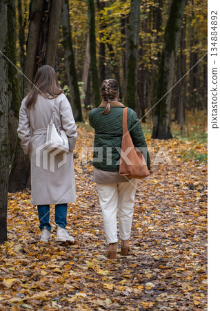 Two women walk away along leaf-covered path among tall trees in Timiryazevsky Park, Moscow, calm and reflective mood Two women walk away along leaf-covered path among tall trees in Timiryazevsky Park, Moscow, calm and reflective mood 134884892