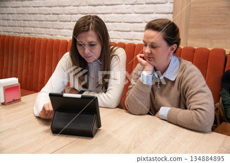 Two women sit in modern cafe using a tablet with electronic menu, focused and relaxed, with warm interior tones and a cozy atmosphere Two women sit in modern cafe using a tablet with electronic menu, focused and relaxed, with warm interior tones and a cozy atmosphere 134884895