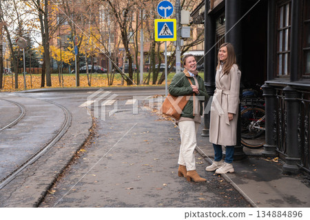 Two women talk and laugh near historic wooden tram stop in Moscow, with wet pavement, tram tracks, street signs, autumn trees in the city background Two women talk and laugh near historic wooden tram stop in Moscow, with wet pavement, tram tracks, street signs, autumn trees in the city background 134884896