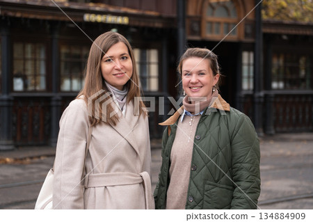Two women walk past a historic wooden tram stop in Moscow, wearing beige and green coats, smiling, with autumn tones and classic urban surroundings 134884909