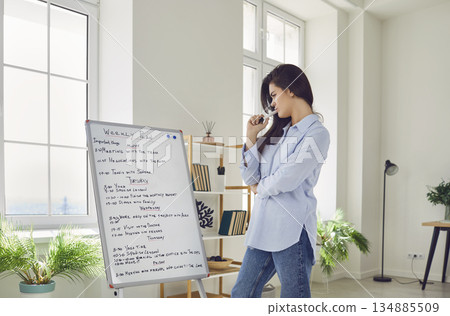 Young business woman standing in office in front of white board with daily plans and tasks Young business woman standing in office in front of white board with daily plans and tasks 134885509