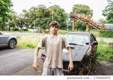 Teenage boy posing confidently beside a wrecked sports car in an industrial urban landscape. Scene expresses masculinity, strength, rebellion and Generation Z street attitude.  134888970