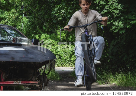 Teenage boy from Generation Z doing a bike trick on a narrow path near an abandoned car, surrounded by green trees. Urban youth lifestyle, freedom, adventure and street culture.  134888972
