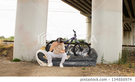 Teenage boy posing with a bicycle on an old sofa under a bridge. Urban lifestyle scene with youth, freedom and creativity in an industrial outdoor environment. Teenage boy posing with a bicycle on an old sofa under a bridge. Urban lifestyle scene with youth, freedom and creativity in an industrial outdoor environment. 134888974