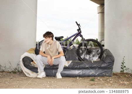 Teenage boy posing with a bicycle on an old sofa under a bridge. Urban lifestyle scene with youth, freedom and creativity in an industrial outdoor environment.  134888975