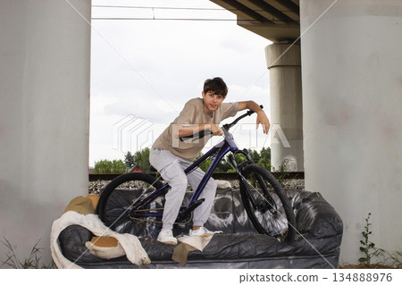 Teenage boy posing with a bicycle on an old sofa under a bridge. Urban lifestyle scene with youth, freedom and creativity in an industrial outdoor environment. Teenage boy posing with a bicycle on an old sofa under a bridge. Urban lifestyle scene with youth, freedom and creativity in an industrial outdoor environment. 134888976