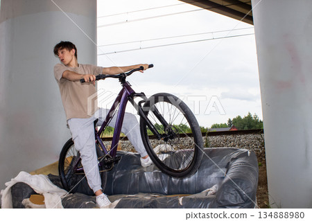 Teenage boy posing with a bicycle on an old sofa under a bridge. Urban lifestyle scene with youth, freedom and creativity in an industrial outdoor environment. Teenage boy posing with a bicycle on an old sofa under a bridge. Urban lifestyle scene with youth, freedom and creativity in an industrial outdoor environment. 134888980