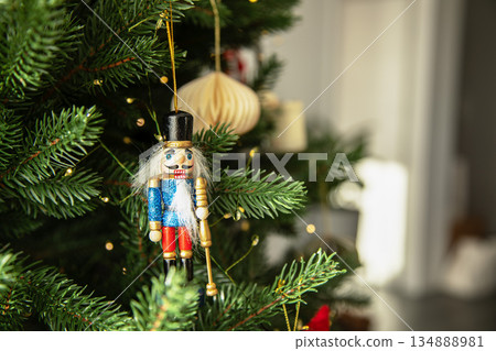 Traditional wooden nutcracker Christmas ornament hanging on a decorated Christmas tree with warm lights and golden baubles. Festive holiday atmosphere with cozy winter mood. 134888981