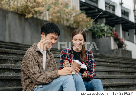 Three young college students is reading a book while relaxing sitting on grass in a campus park with her friends. 134889627