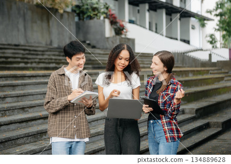 Three young college students is reading a book while relaxing sitting on grass in a campus park with her friends. 134889628