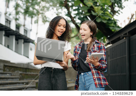 Three young college students is reading a book while relaxing sitting on grass in a campus park with her friends. 134889629