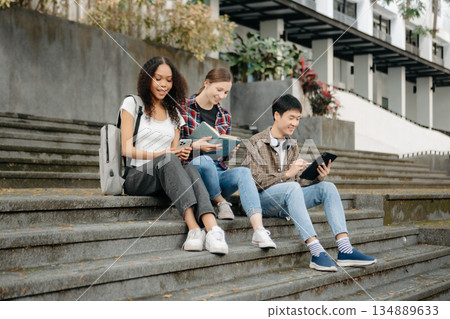 Three young college students is reading a book while relaxing sitting on grass in a campus park with her friends. 134889633